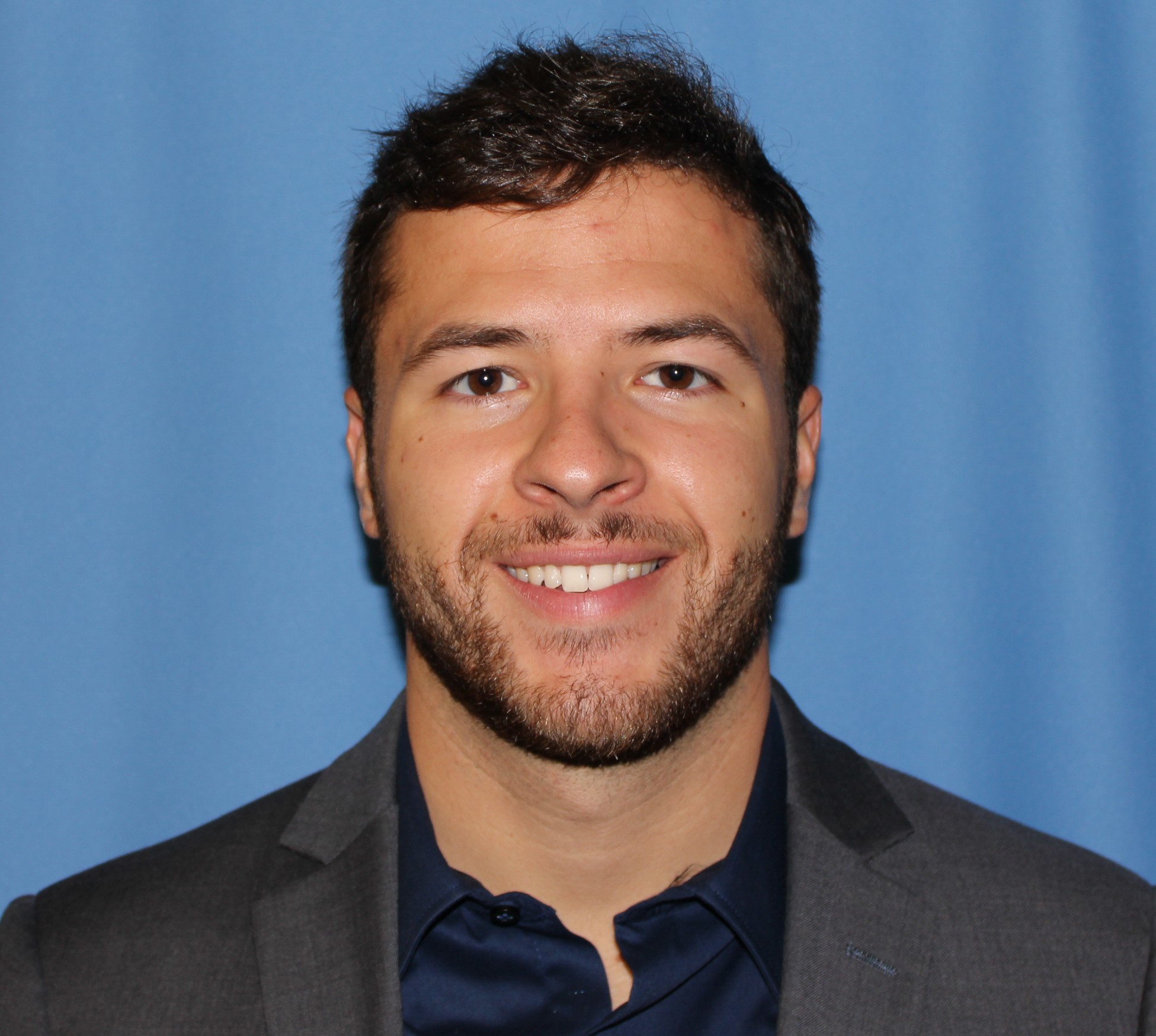 MVSS Benefits Coordinator Joe Galeaz smiles for a headshot photo against a blue background wearing a grey jacket.